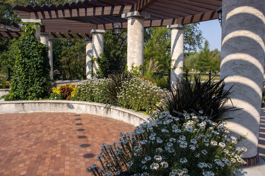 A slatted timber shade structure on masonry columns casting striped shade across a paver field, with a curved seating wall doubling as a raised planter